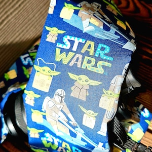 May include: Blue fabric featuring Star Wars characters, including The Mandalorian and Grogu. The words "STAR WARS" are printed in a colourful font. The fabric is wrapped around a black object.