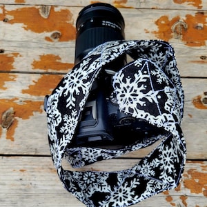 May include: A black camera strap with a white snowflake pattern. The strap is wrapped around a black camera lens and body.
