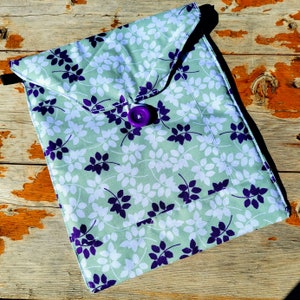 Puede incluir: Un sobre de tela verde con cierre de botón morado. La tela tiene un patrón de hojas blancas y moradas.