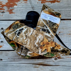 May include: A camouflage camera strap with the text "Moorewood Designs" and "moorewooddesigns.com" printed on it. The strap is wrapped around a black camera lens.