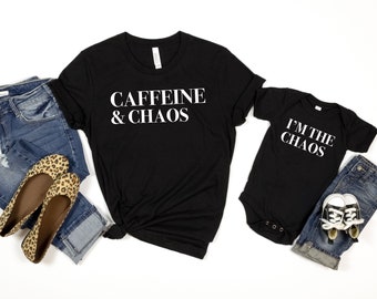 May include: A black t-shirt with the text "CAFFEINE & CHAOS" in white lettering. A matching black baby onesie with the text "I'M THE CHAOS" in white lettering. The shirts are laid out on a white background with a pair of jeans and leopard print flats.