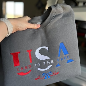 May include: A gray sweatshirt with the embroidered text "USA LAND OF THE FREE" in red, white, and blue. The design includes a bow and decorative elements in the same colors, celebrating American patriotism.