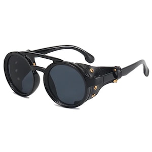 May include: Black round sunglasses with dark lenses and a distinctive steampunk aesthetic. The sunglasses have a black frame, leather side shields, and gold rivets. The arms curve for a comfortable fit.
