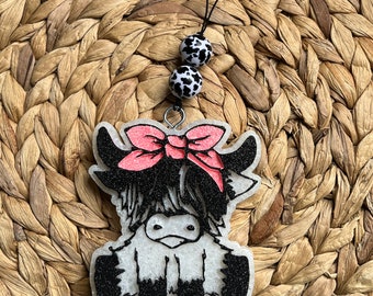 May include: A white Highland cow charm with black and white spots, wearing a pink bow, hanging from a black cord with black and white spotted beads.