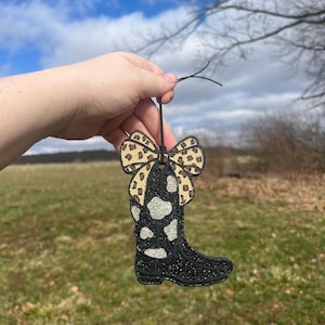 May include: A black glitter boot with a white and black cow print design. The boot has a leopard print bow on top.