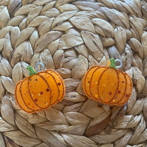 May include: Two orange pumpkin-shaped earrings with glitter and green stems. The earrings are set against a woven, beige background. The pumpkins have a textured appearance and are decorated with small, dark orange and brown spots.
