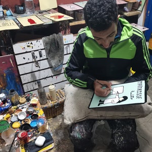 May include: A person is painting a sign with a black marker. The sign is green and white and says "Maitrisse" and "aliz&eacute;" in black lettering. The sign also has a black and white illustration of a person wearing a white coat and holding a paintbrush.