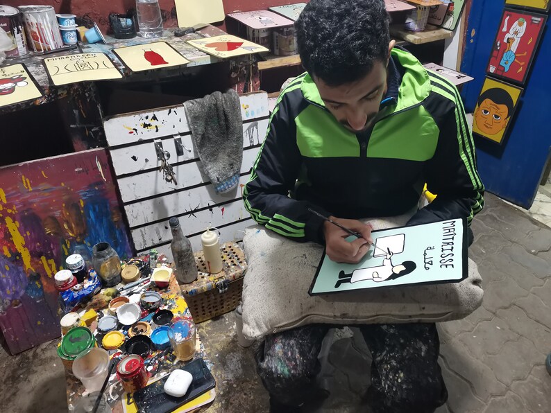 May include: A person is painting a sign with a black marker. The sign is green and white and says "Maitrisse" and "aliz&eacute;" in black lettering. The sign also has a black and white illustration of a person wearing a white coat and holding a paintbrush.