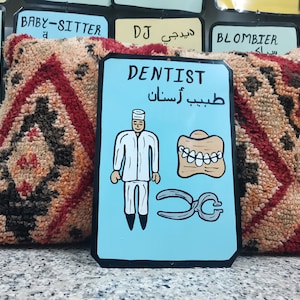May include: A blue and white sign with the word "DENTIST" in black lettering. The sign also includes a cartoon image of a dentist, a set of teeth, and a pair of pliers. The text "طبيب أسنان" is written in Arabic below the word "DENTIST".