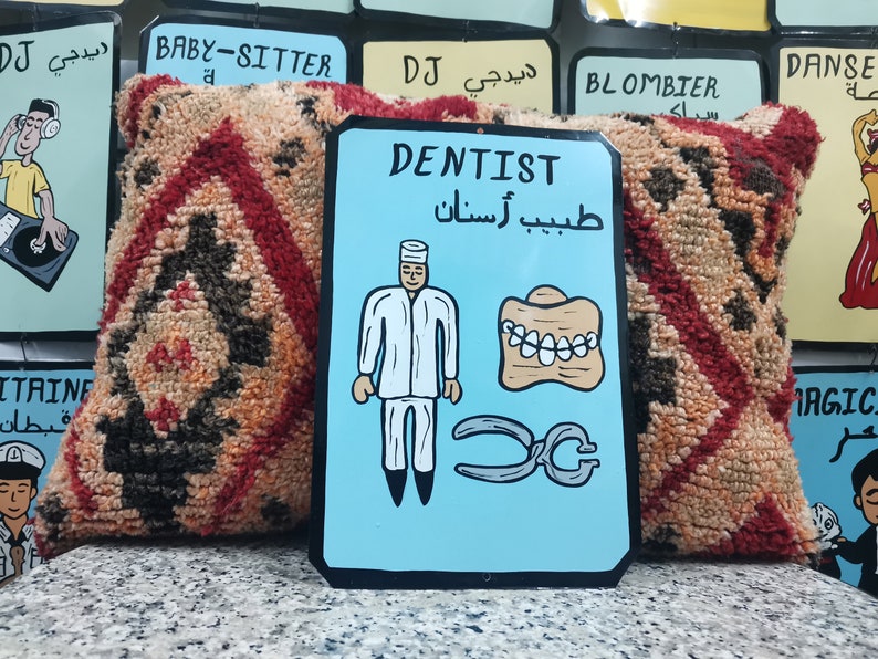 May include: A blue and white sign with the word "DENTIST" in black lettering. The sign also includes a cartoon image of a dentist, a set of teeth, and a pair of pliers. The text "طبيب أسنان" is written in Arabic below the word "DENTIST".
