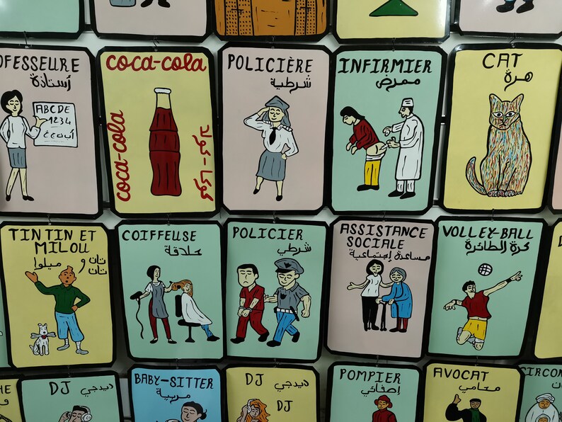 May include: A colorful wall display of illustrated cards with various professions and activities. Each card features a cartoon image and text in both Arabic and French. The professions include teacher, police officer, nurse, hairdresser, social worker, volleyball player, DJ, babysitter, firefighter, and lawyer.