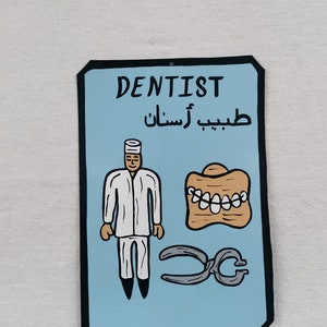 May include: A blue and black sign with the word "DENTIST" in white lettering. The sign also includes the Arabic text "طبيب أسنان" and a cartoon illustration of a dentist, a set of teeth, and a pair of pliers.