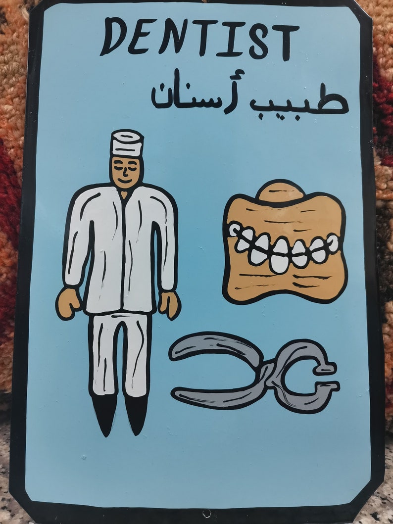 May include: A blue and white sign with the word "DENTIST" in black lettering. The sign also includes the Arabic word for dentist, "طبيب أسنان", in black lettering. The sign features a cartoon illustration of a dentist, a set of teeth, and a pair of pliers.
