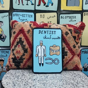 May include: A blue and white sign with the word "DENTIST" in English and "طبيب أسنان" in Arabic. The sign features a cartoon dentist wearing a white coat and holding a pair of pliers. There are also cartoon teeth and a set of dentures on the sign.