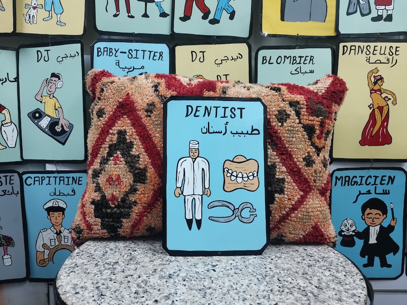 May include: A blue and white sign with the word "DENTIST" in English and "طبيب أسنان" in Arabic. The sign features a cartoon dentist wearing a white coat and holding a pair of pliers. There are also cartoon teeth and a set of dentures on the sign.