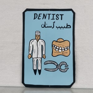 May include: A blue and black sign with the word "Dentist" in white lettering. The sign also includes a cartoon image of a dentist, a set of dentures, and a pair of pliers. The Arabic text on the sign reads "طبيب أسنان".