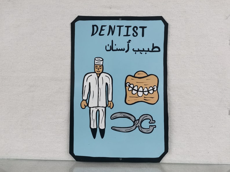 May include: A blue and black sign with the word "Dentist" in white lettering. The sign also includes a cartoon image of a dentist, a set of dentures, and a pair of pliers. The Arabic text on the sign reads "طبيب أسنان".