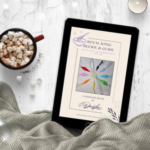 May include: A digital tablet displays a recipe guide for royal icing, with colorful icing designs. A mug of hot chocolate with marshmallows sits next to the tablet. Two candles and a gray blanket complete the scene.
