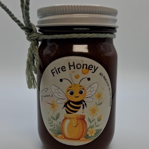 May include: A jar of Fire Honey with a white lid and a label featuring a cartoon bee and a honey pot. The jar is tied with a green string. The label reads "Fire Honey" and "All Natural - So Good."
