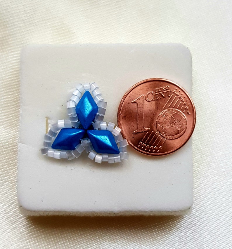 May include: A small blue and white beaded flower design, approximately the size of a 1 Euro cent coin.