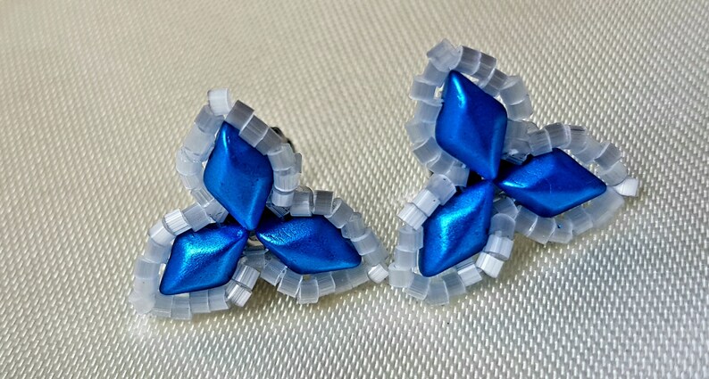 May include: A pair of blue and white beaded earrings in the shape of a three-leaf clover. The earrings are made with small, round beads and have a shiny, metallic finish.