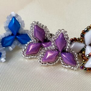 May include: Three pairs of beaded earrings in a flower shape. The first pair is blue and white, the second pair is purple, and the third pair is white with iridescent accents.
