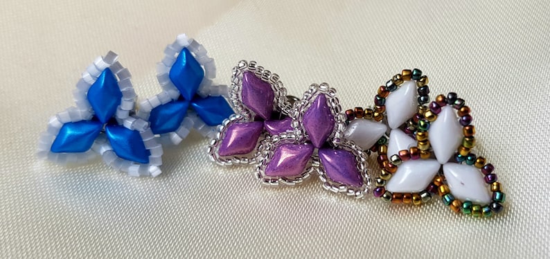 May include: Three pairs of beaded earrings in a flower shape. The first pair is blue and white, the second pair is purple, and the third pair is white with iridescent accents.