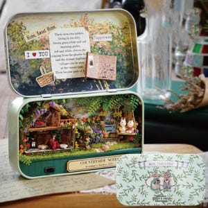 Può includere: Una scena di casa delle bambole in miniatura all'interno di una scatola di metallo verde. La scena presenta una piccola casa di legno con un'altalena sul portico, un giardino con fiori e alberi, e due conigli bianchi seduti sull'altalena. Il coperchio della scatola presenta una scena dipinta di un giardino con una citazione sulla felicità. La scatola è etichettata "NOTE CAMPESTRE Giardino della nonna, 1883" e "TEATRO DI SCATOLA".