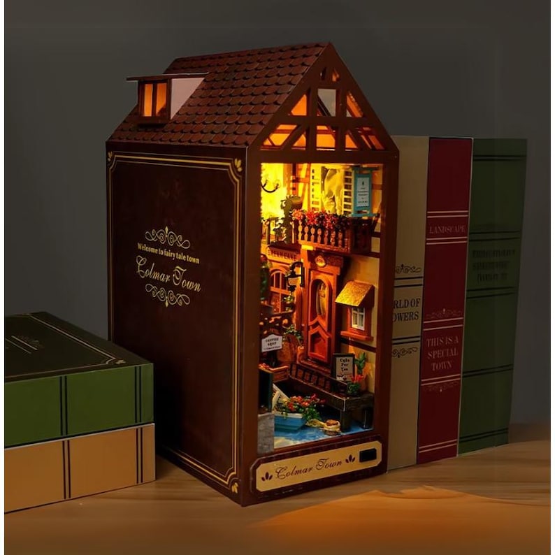 May include: A detailed, illuminated diorama of a town scene, housed within a book-shaped structure. The brown exterior features the text "Welcome to fairy tale town Colmar Town". The interior shows a street with balconies and flowers. Books are to the right.