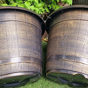 May include: Two large, round, dark brown planters with a textured, wood-grain appearance and gold accents. The planters have a wide rim and a black base, suitable for outdoor use. They are sitting on green grass.