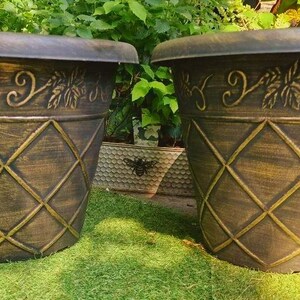 May include: Two matching tapered planters with a dark bronze finish and a gold-toned lattice pattern on the lower body. The upper rim features a raised relief design of scrolling vines and leaves, creating a textured appearance on the exterior.