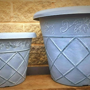 May include: Two light blue decorative planters. The planters have a diamond pattern and a decorative leaf and vine design around the top. The planters are made of a durable material and are suitable for indoor or outdoor use.