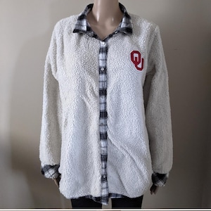 May include: A white, fuzzy, button-down shirt with a black and white plaid collar, cuffs, and inner placket. The shirt features a red and white "OU" patch on the chest. The shirt is displayed on a mannequin.