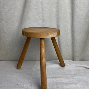 Può includere: Uno sgabello di legno semplice con tre gambe e una seduta rotonda. Lo sgabello è realizzato in legno marrone chiaro e ha un aspetto rustico.