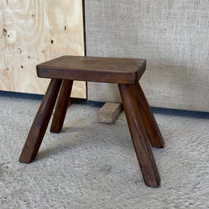 Op de afbeelding: Een kleine, handgemaakte houten kruk met een vierkante zitting en drie schuine poten. De kruk is gemaakt van donkerbruin hout en heeft een rustiek, minimalistisch ontwerp. De kruk is ongeveer 30 cm hoog.