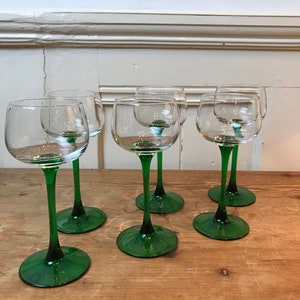 Può includere: Sei calici da vino trasparenti con steli e basi verdi sono disposti su una superficie di legno. I calici hanno un design classico, con una coppa arrotondata e un gambo lungo e sottile. Il colore verde degli steli e delle basi aggiunge eleganza.