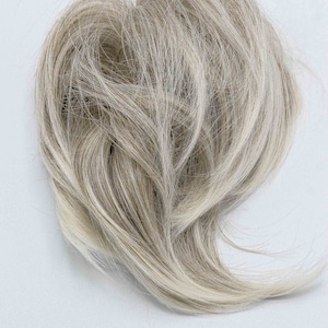 May include: A light-colored hairpiece, likely a wig or hair extension, with a blend of blonde and gray strands. The hair is styled in a loose, wavy pattern, creating a natural look. The hairpiece is set against a white background.
