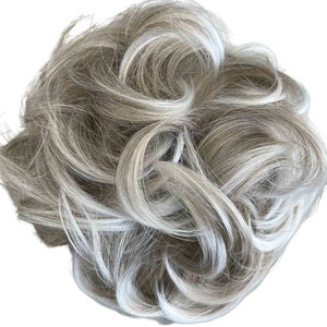 May include: A grey and white hair bun with a curly texture. The hairpiece is a mix of light grey and white strands, creating a natural, blended look. The bun is designed to be worn as a hair accessory.
