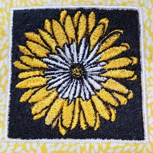 Könnte beinhalten: Ein gestickter Blumen-Patch in Gelb und Weiß mit schwarzem Hintergrund. Die Blume hat eine weiße Mitte und gelbe Blütenblätter.