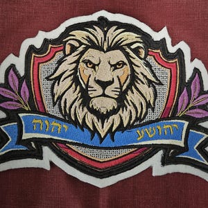 Peut inclure: Un écusson brodé représentant une tête de lion dans un bouclier avec une bannière qui dit "דרושי" et "לורה". Le lion est beige et brun avec un contour noir. Le bouclier est rouge, blanc et bleu avec un contour noir. La bannière est bleue avec un contour noir et des lettres dorées.
