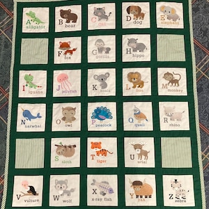 May include: A handmade alphabet quilt featuring embroidered animals and their corresponding letters. The quilt showcases a green border and white squares with various animals like a bear, cat, dog, and zebra. The quilt is designed for educational purposes.