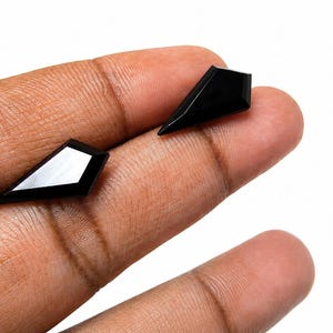 May include: Two loose, black, kite-shaped gemstones with a faceted surface. Each stone features a sharp, pointed geometry with a smooth, reflective finish, showcasing a deep, solid black color and precise, angular edges on a white background.