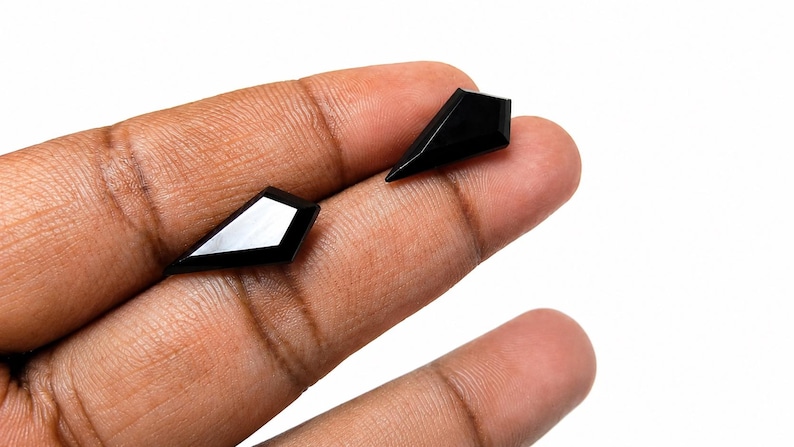 May include: Two loose, black, kite-shaped gemstones with a faceted surface. Each stone features a sharp, pointed geometry with a smooth, reflective finish, showcasing a deep, solid black color and precise, angular edges on a white background.