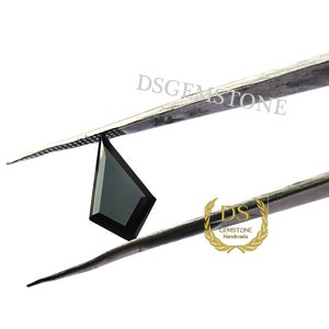 May include: A kite-shaped black gemstone held by metal tweezers. The stone features a faceted surface with sharp, geometric edges. The text DSGEMSTONE is visible above the stone, and a gold logo with the text DS GEMSTONE Handmade is below.