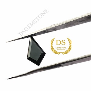May include: A kite-shaped black gemstone held by metal tweezers. The stone has a faceted surface and a dark, opaque appearance. The text DSGEMSTONE is visible along the top tweezer, and a gold logo with the text DS GEMSTONE Handmade is present.