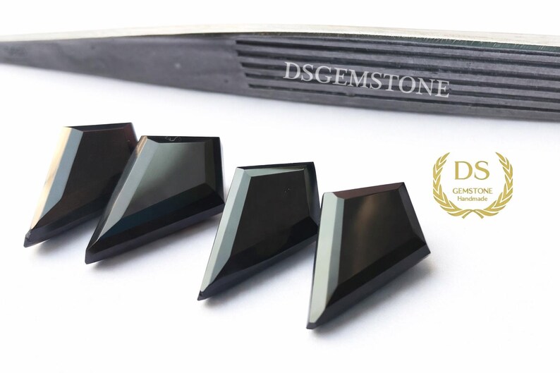 May include: Four black shield-shaped faceted gemstones arranged in a row. Above the stones is a pair of dark metal tweezers with the text DSGEMSTONE engraved. To the right is a gold laurel wreath logo with the text DS GEMSTONE Handmade.