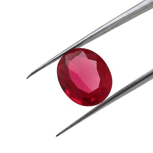 May include: A faceted, oval-shaped ruby gemstone held by silver tweezers. The ruby is a deep, vibrant red colour, and the tweezers are positioned against a white background.
