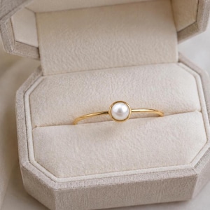 Könnte beinhalten: Ein dünner Goldring mit einer einzelnen, kleinen, runden weißen Perle in einer Zargenfassung. Der Ring liegt horizontal in einer hellbeigen, achteckigen Schmuckschatulle aus Samt mit einer weich gepolsterten Innenauskleidung.