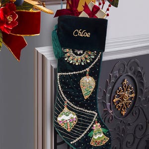 May include: A dark green velvet Christmas stocking featuring the name Chloe embroidered in gold thread at the top. The stocking is decorated with intricate beadwork and sequins in patterns of hanging ornaments and draped chains in multi-colored tones.