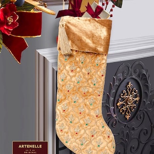 Puede incluir: Un calcetín navideño de terciopelo dorado con un patrón de celosía repetido bordado con hilo de oro, acentuado por pequeñas cuentas verdes y rojas. El calcetín incluye una borla dorada en el lateral y está lleno de dos cajas de regalo con cintas burdeos.